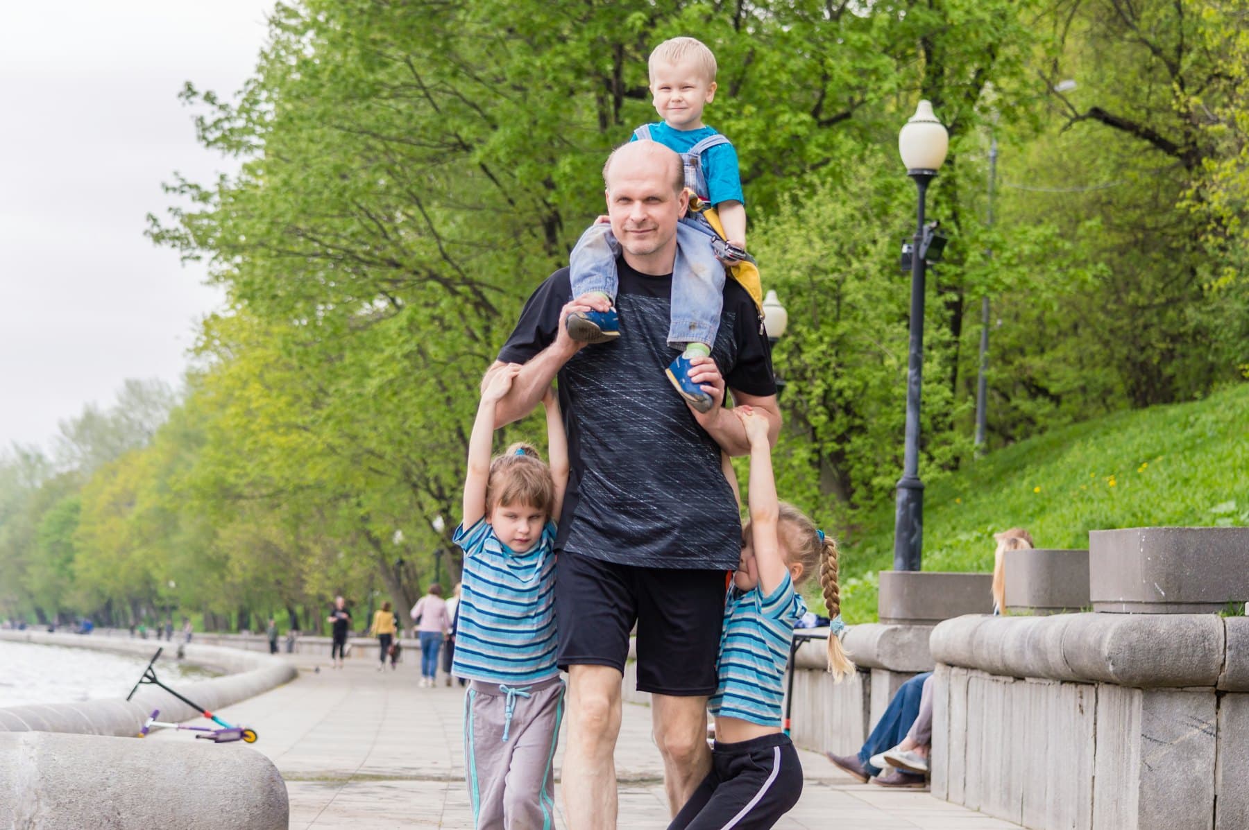 A father walking in the park with his kids, one riding on his shoulders
