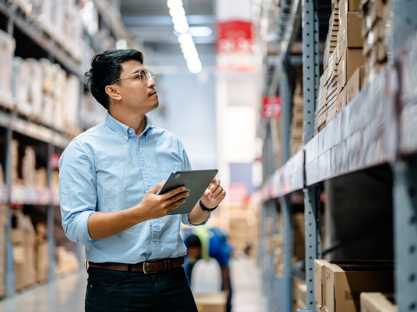 A warehouse manager checking stock on a tablet between tall shelves of boxes