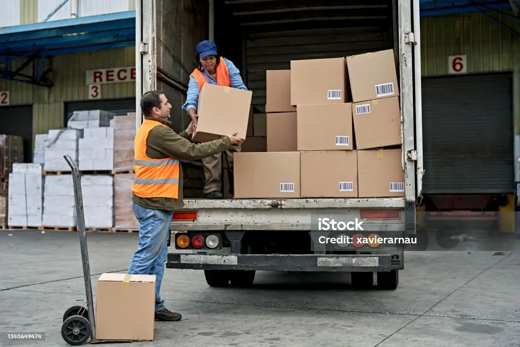 Two workers load barcoded boxes into a truck at a distribution center dock.
